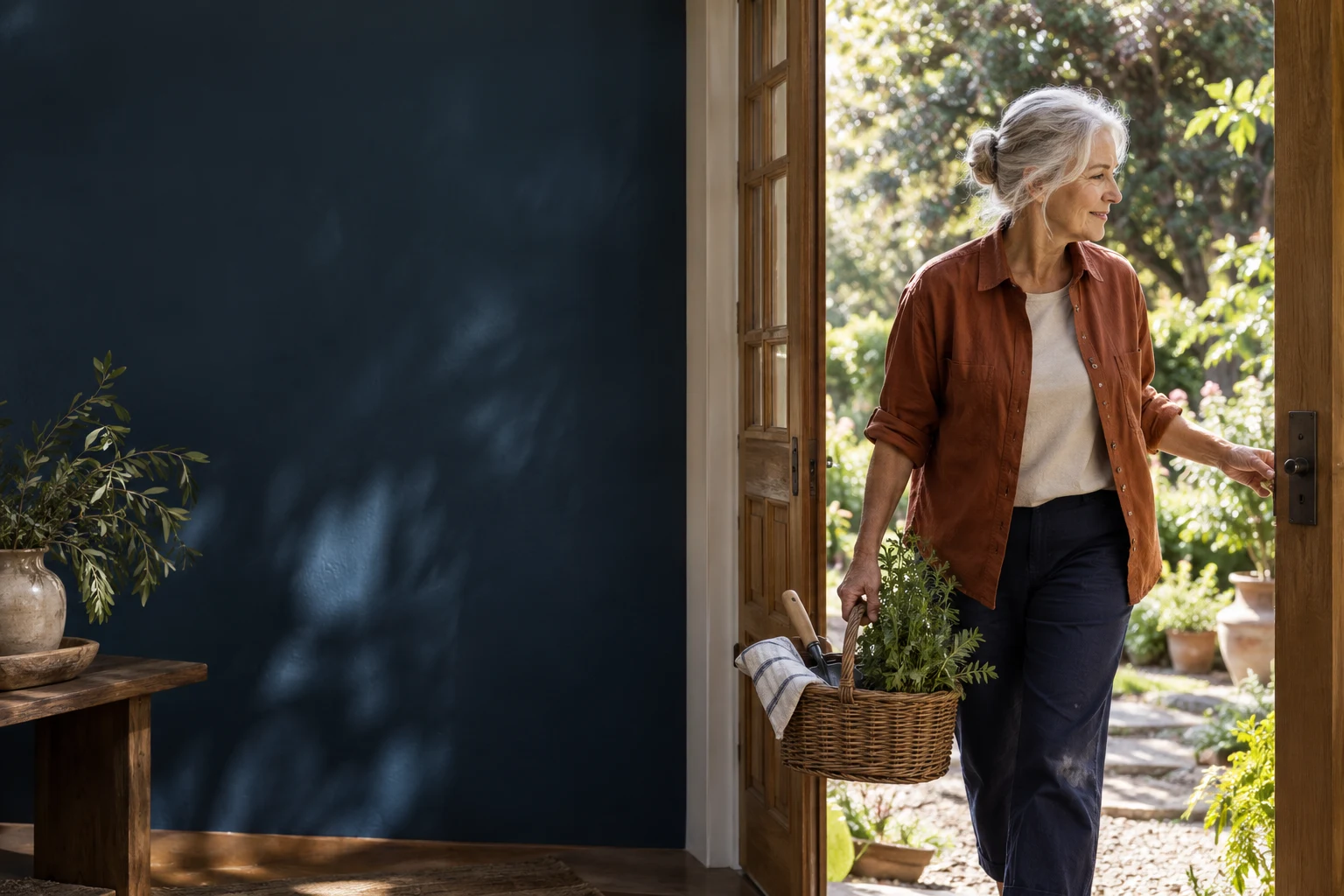 A silver-haired woman carrying a basket of garden tools toward her sunlit garden
